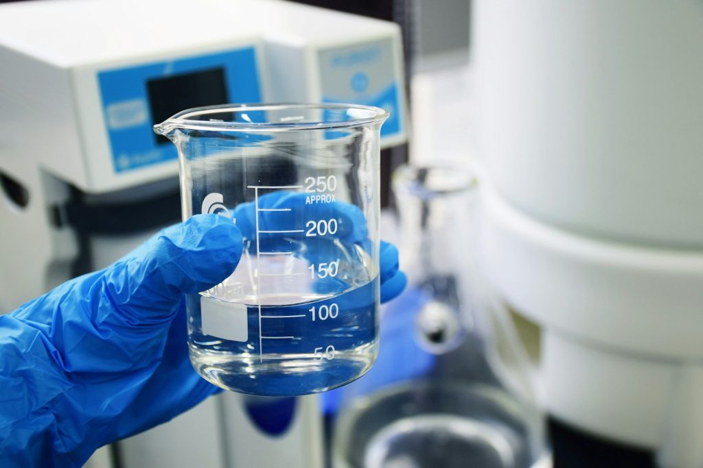 A person wearing blue nitrile gloves holding a glass beaker of water in a laboratory setting.